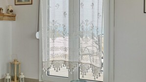 A room with a glass table, a white door with a lace curtain, and a small shelf with decorative items. - A BALCONY WITH AMAZING SEA VIEW (CHIOS)