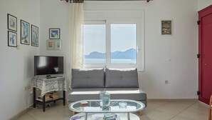 A living room with a sofa, a TV, a glass coffee table, and a view of mountains through a window. - A BALCONY WITH AMAZING SEA VIEW (CHIOS)