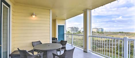 Un patio cubierto con una mesa redonda y sillas de mimbre, una puerta azul y vista a la playa y edificios en la distancia.