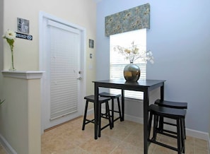 A room with a white door, a window with blinds, a vase with flowers, and a table with two stools.