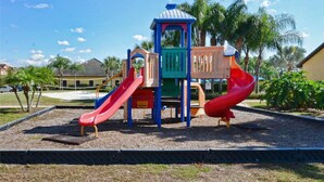 Ein Spielplatz mit zwei Rutschen und einer Kletterkonstruktion, umgeben von Palmen und einem Gebäude im Hintergrund.