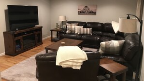 Uma sala de estar com sofá de couro, mesa de centro, televisão sobre suporte de madeira, abajur e um quadro na parede.