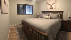 Quarto com cama de madeira, duas mesas de cabeceira com luminárias, janela com persianas e quadro de um veado na parede.