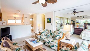 A spacious living room with a large sofa, a coffee table, and a television. There is a kitchen area with a refrigerator and a dining table. The room has a ceiling fan and a large window. - Tropical Family Pick w/Kitchen Ease, Free WiFi, Washer/Dryer–Waikīkī Shore 318 (HONOLULU)