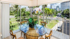 A corner room with a glass table, four wicker chairs, a large window with a view of palm trees and city buildings. - Tropical Family Pick w/Kitchen Ease, Free WiFi, Washer/Dryer–Waikīkī Shore 318 (HONOLULU)