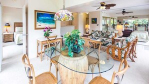 A spacious living area with a glass dining table surrounded by wicker chairs, a large painting of a beach scene, and a ceiling fan. - Tropical Family Pick w/Kitchen Ease, Free WiFi, Washer/Dryer–Waikīkī Shore 318 (HONOLULU)