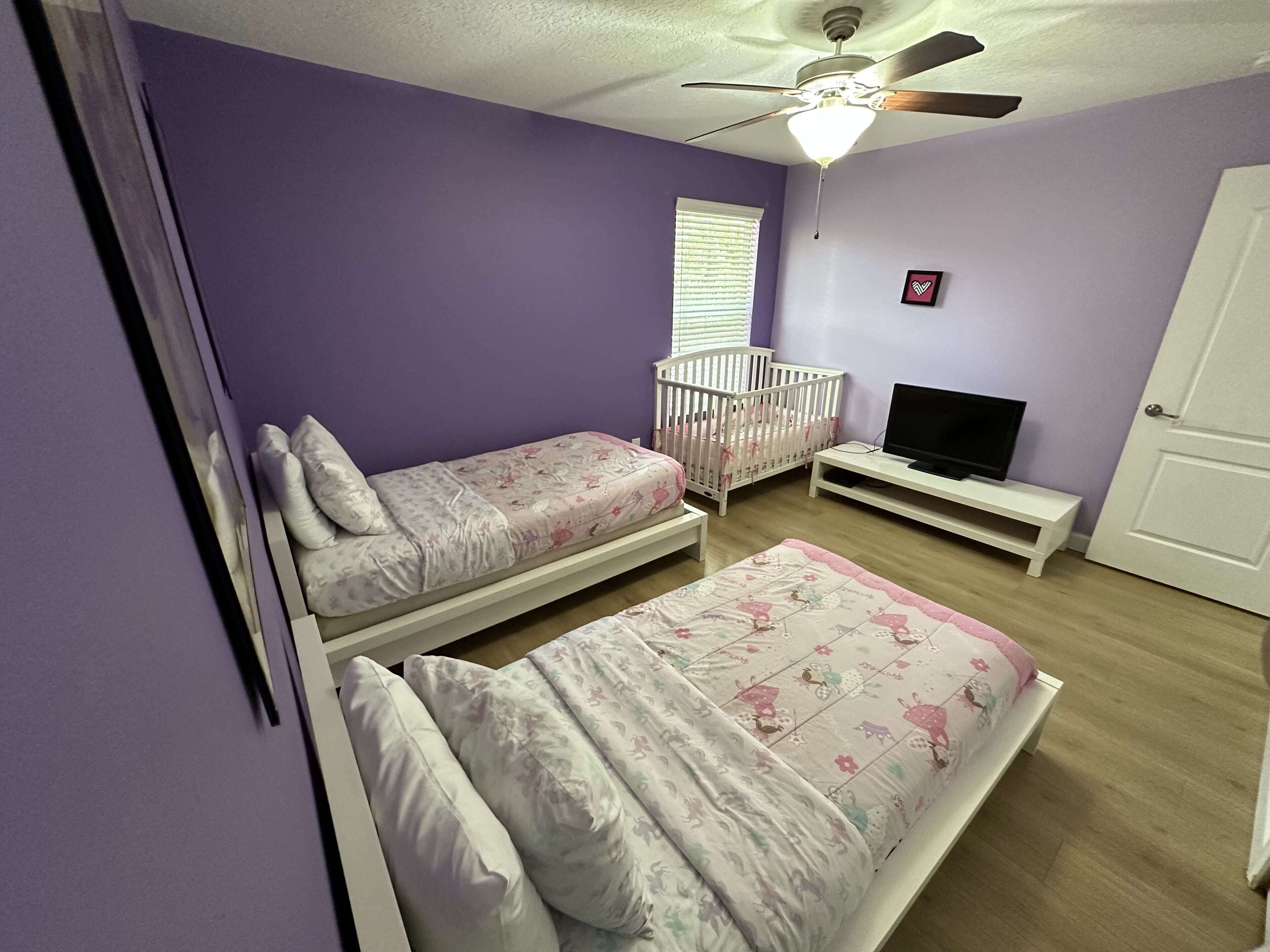 A bedroom with two single beds, a crib, a television on a stand, and a ceiling fan.