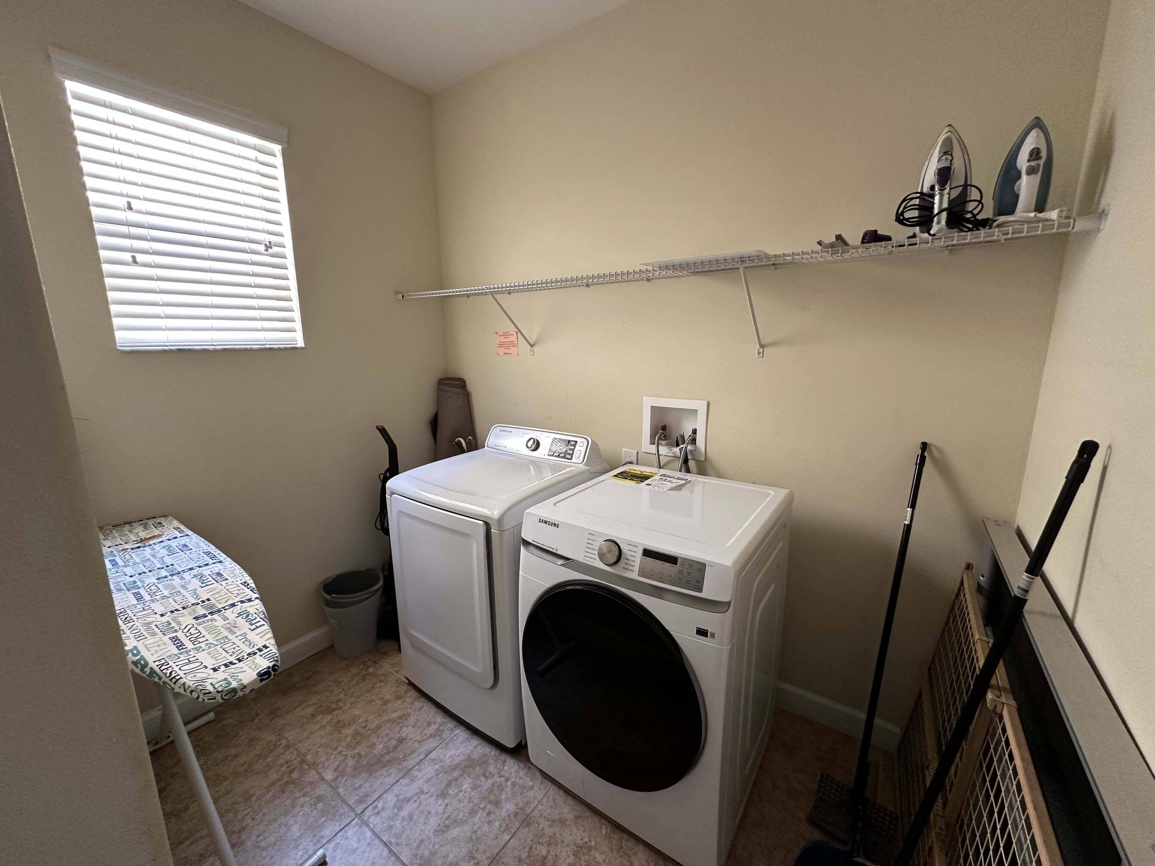 A laundry room with a washing machine, dryer, ironing board, and shelves with ironing boards.