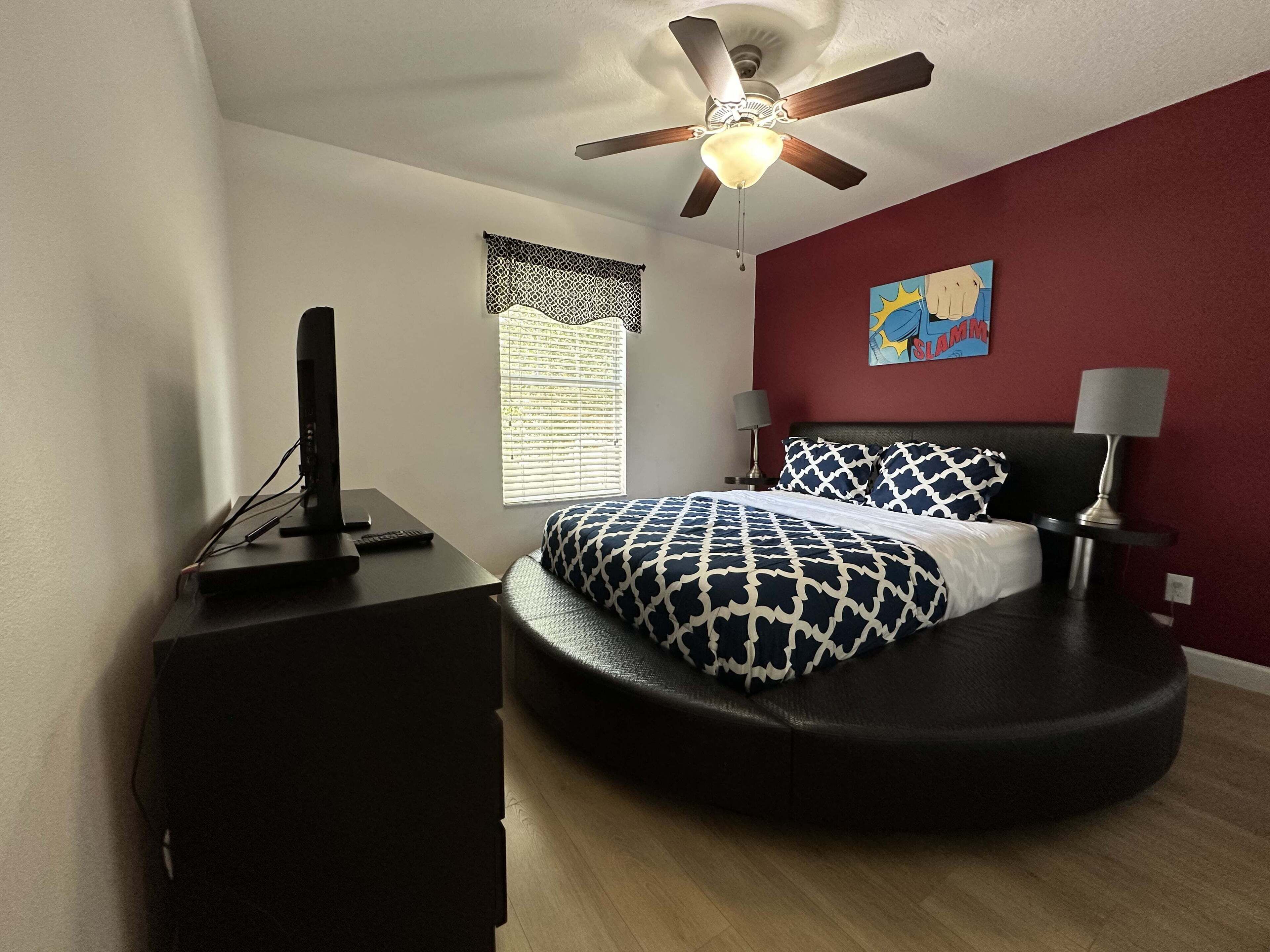 A bedroom with a bed, a ceiling fan, a window with blinds, a painting on the wall, and a desk with a lamp.