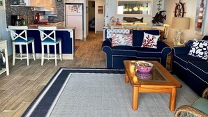 Sala de estar com sofá azul, mesa de centro e área de cozinha com geladeira e banquetas.