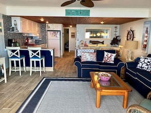 A living room with a blue sofa, a coffee table, and a kitchen area with a refrigerator and bar stools. - Relaxing, Stylish 1 Bedroom Oceanfront Condo with Free WiFi, an Outdoor Pool, Beach Decor, and a Great Ocean View Located in Midtown and Right by Beach! (Ocean City)