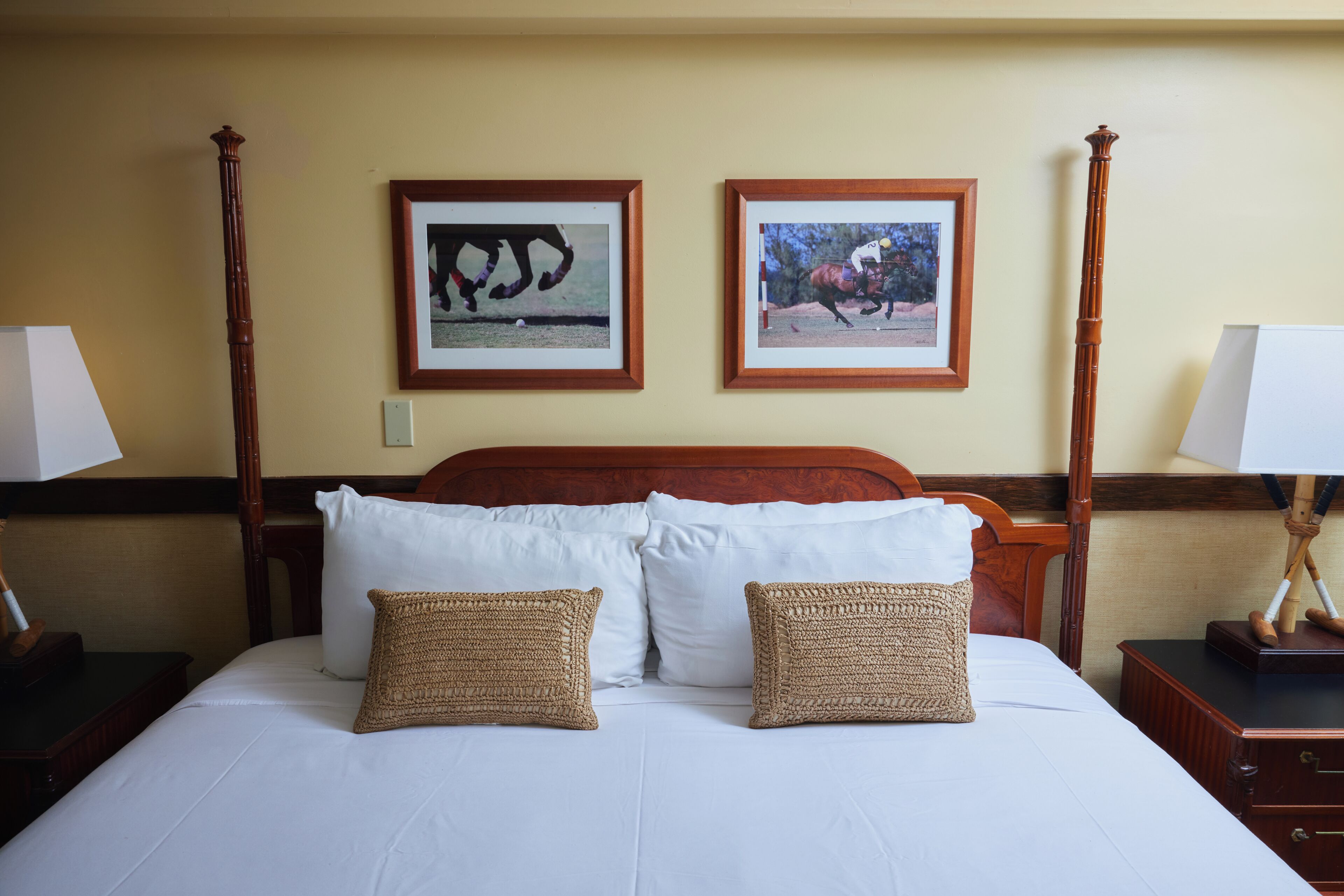 A hotel room with a wooden bed, white linens, two framed horse racing photographs, and a nightstand with a lamp.