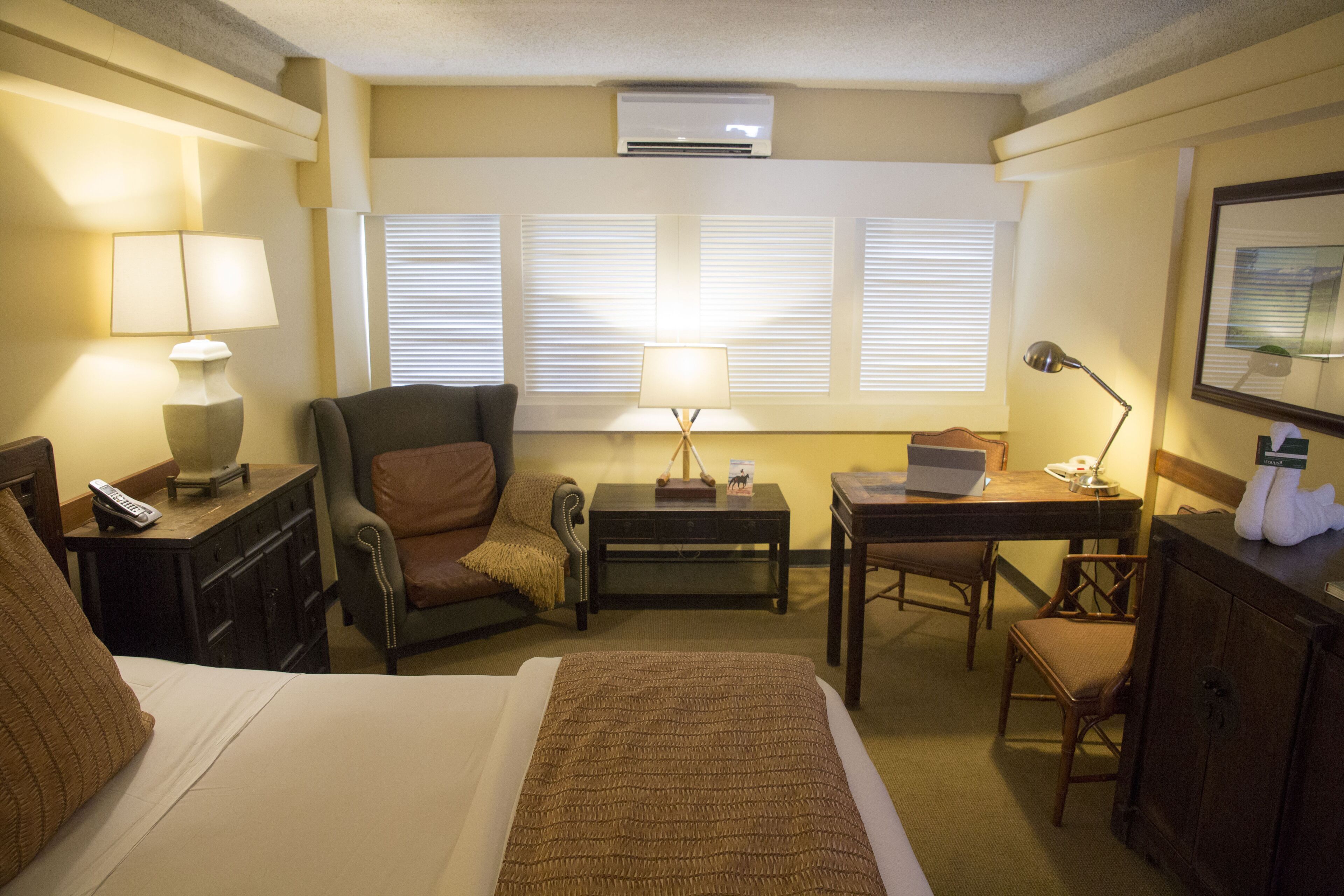 Une chambre d’hôtel avec un lit, un bureau, une chaise, une lampe et une fenêtre avec des stores.