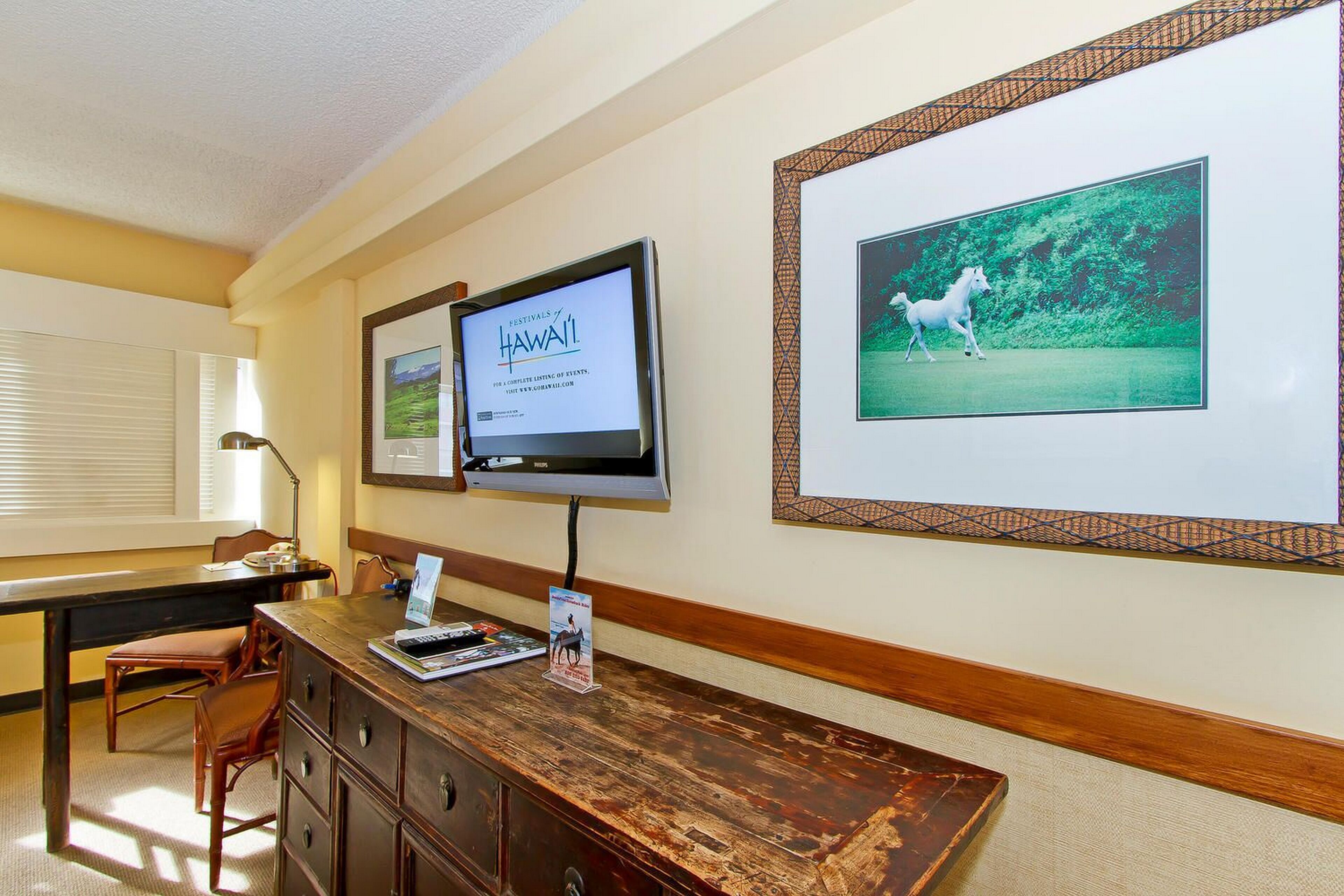 A reception desk with a television displaying 'Festivals of Hawaii', a framed picture of a horse, and a wooden desk with drawers.