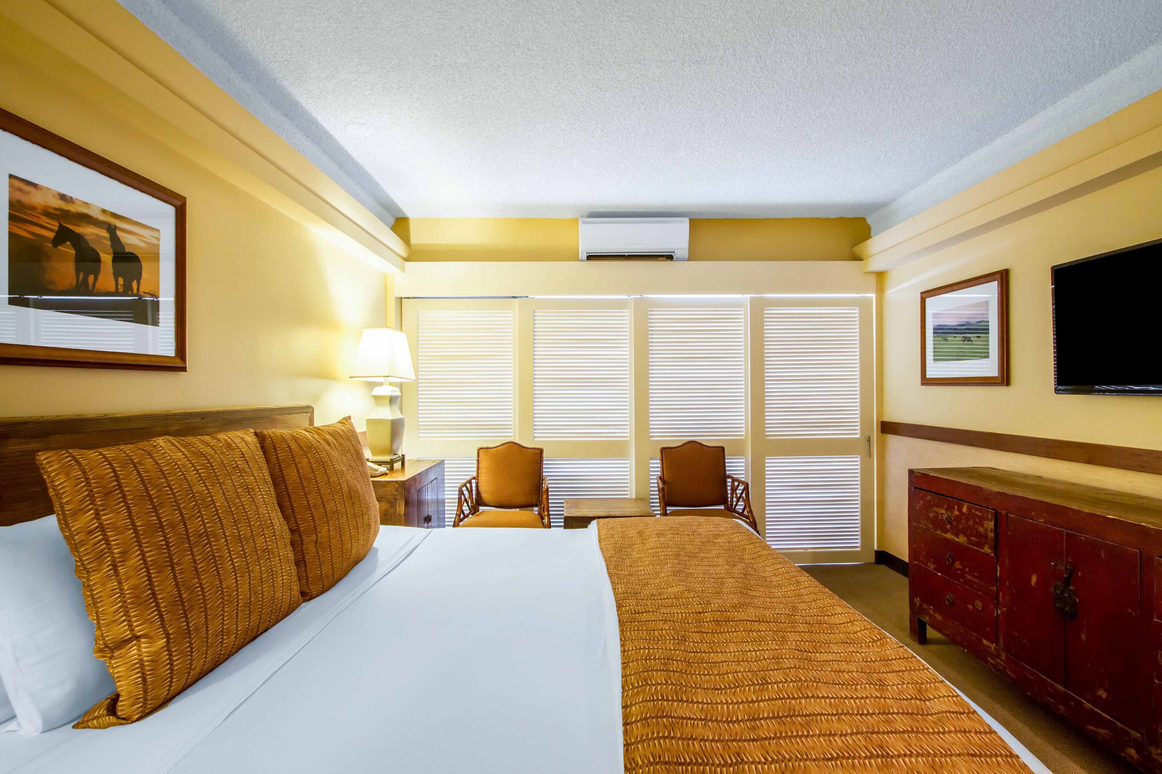 Habitación de hotel con una cama grande, dos sillas, un aparador de madera y un televisor en la pared.