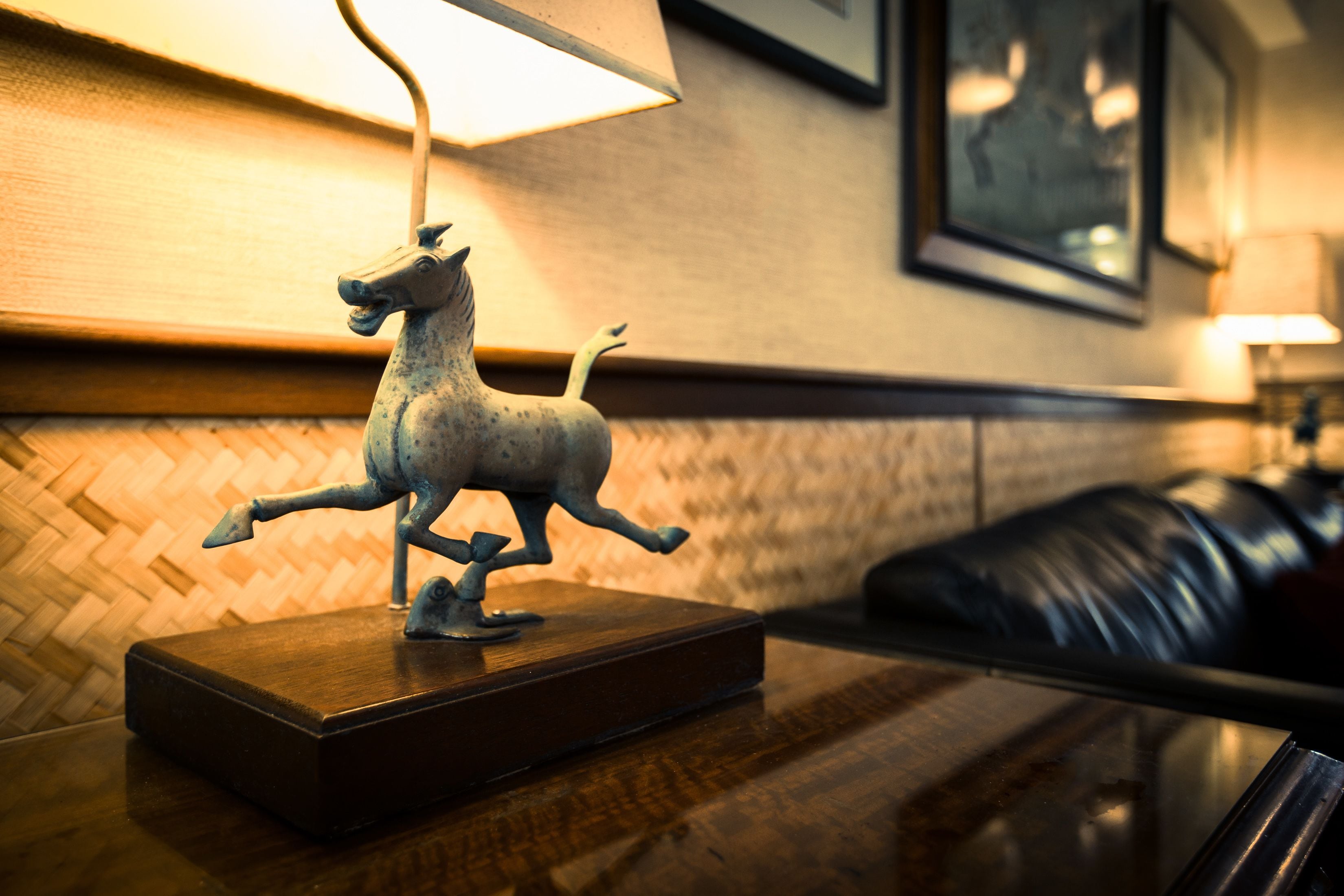 A small bronze horse figurine on a wooden base, a lamp, and a black leather couch in a warmly lit room with a woven wall pattern.