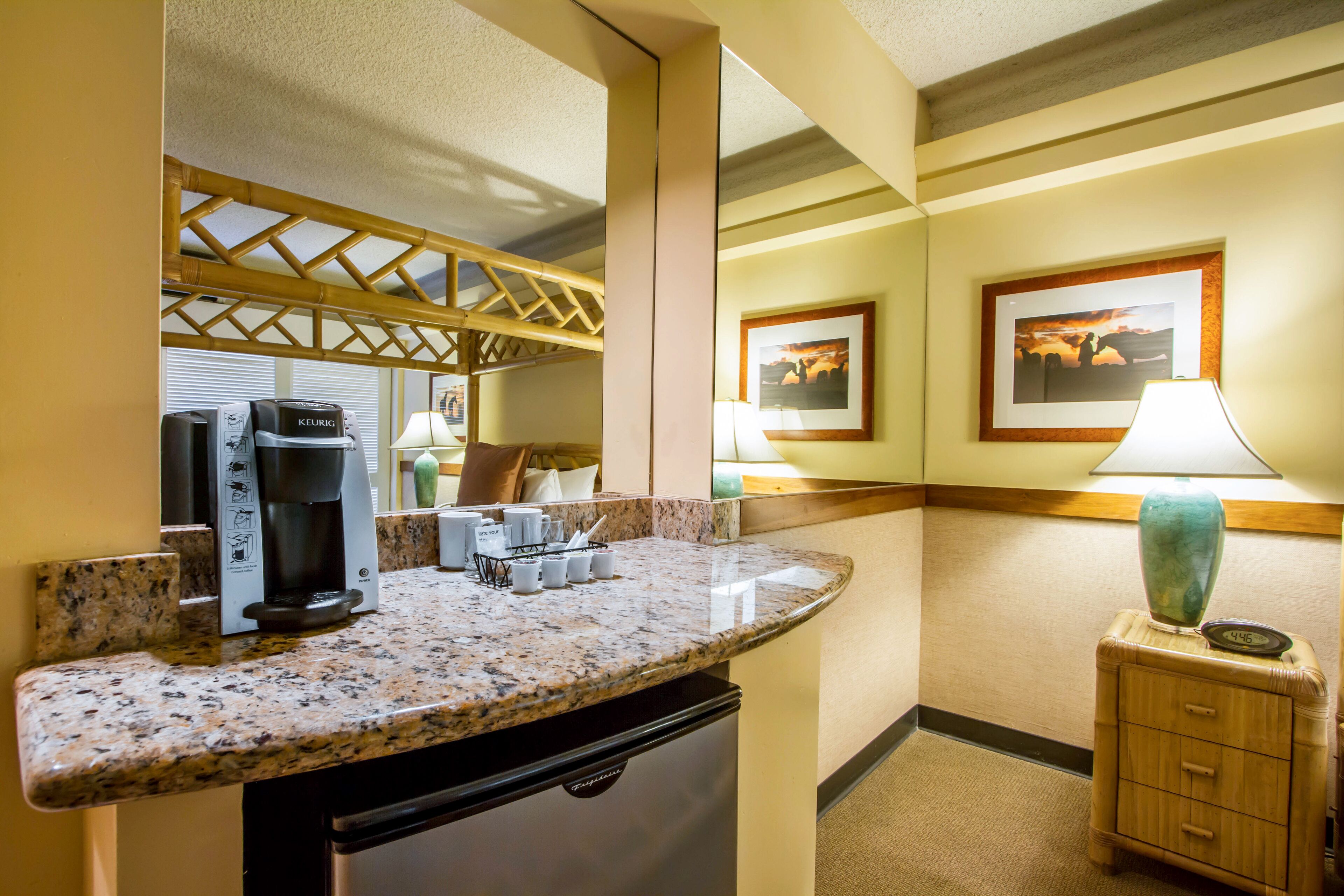 A hotel room with a kitchenette, a coffee maker, a lamp, and framed pictures on the wall.