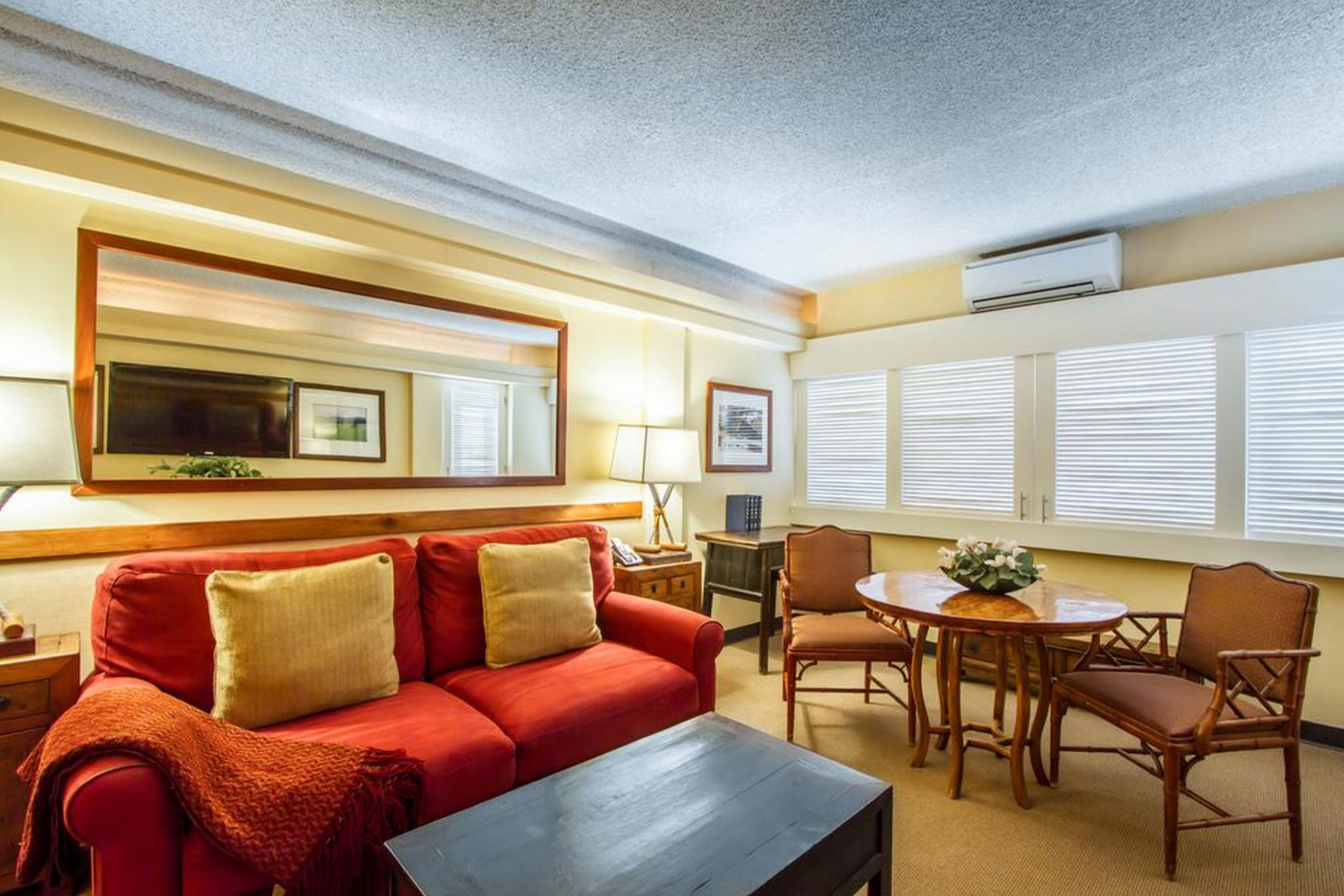 Une chambre d’hôtel avec un canapé rouge, une table basse en bois, une table de salle à manger ronde avec des chaises, un grand miroir et un climatiseur fixé au mur.