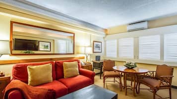Une chambre d’hôtel avec un canapé rouge, une table basse en bois, une table de salle à manger ronde avec des chaises, un grand miroir et un climatiseur fixé au mur.