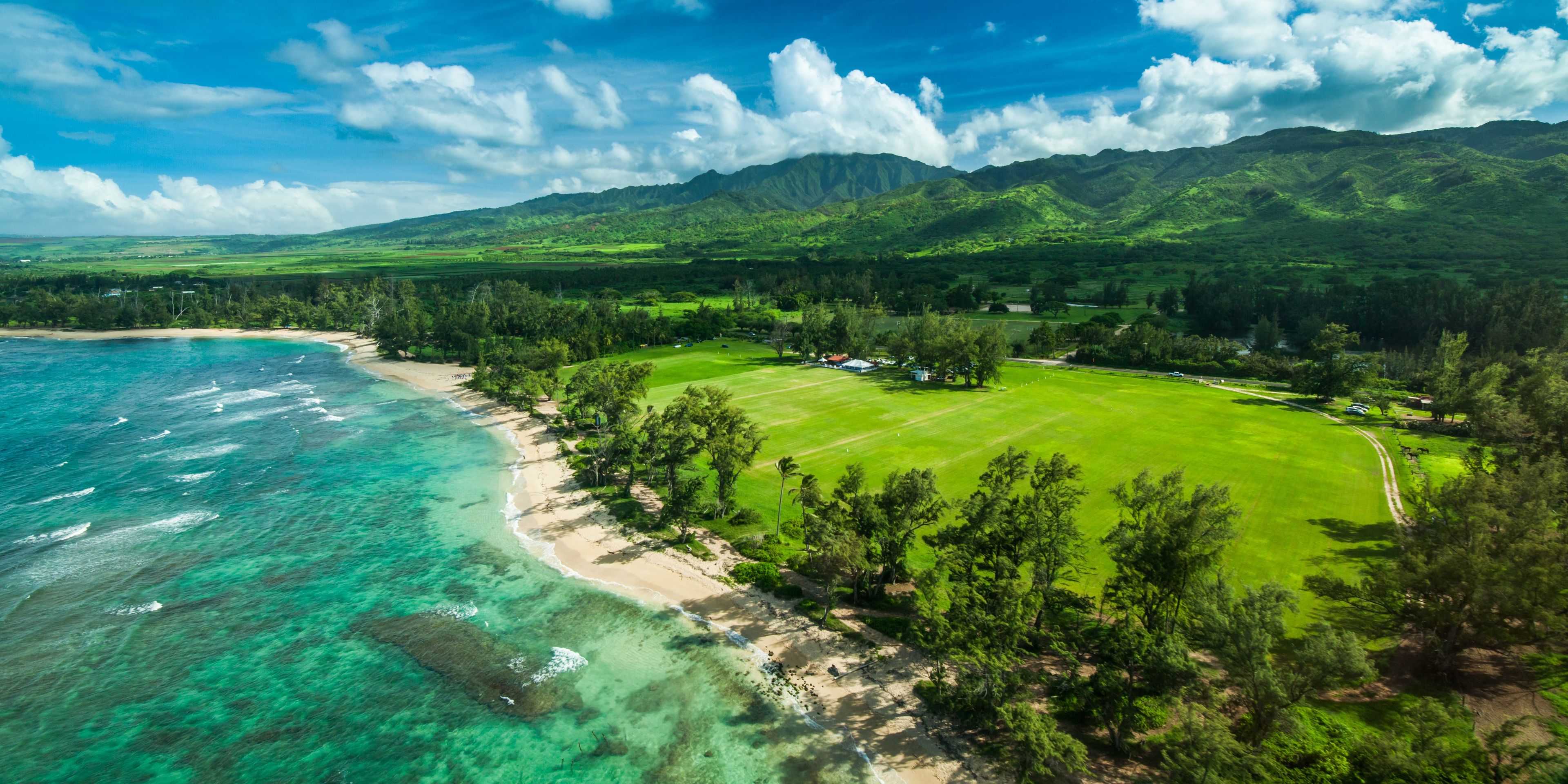 Un paisaje costero con una playa, aguas turquesas cristalinas y un campo de golf rodeado de colinas verdes exuberantes.