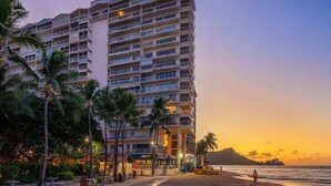 Een hotel met meerdere verdiepingen en balkons, nabij een strand met palmbomen en uitzicht op een zonsondergang.