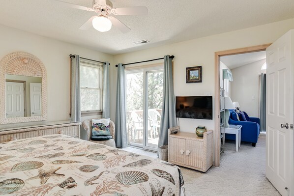 A bedroom with a bed, a ceiling fan, a television, a wicker dresser, a blue sofa, and a sliding glass door leading to a balcony. - My Happy Place: Private Pool & Hot Tub, Rec Room w/ Foosball and Air Hockey (Corolla)