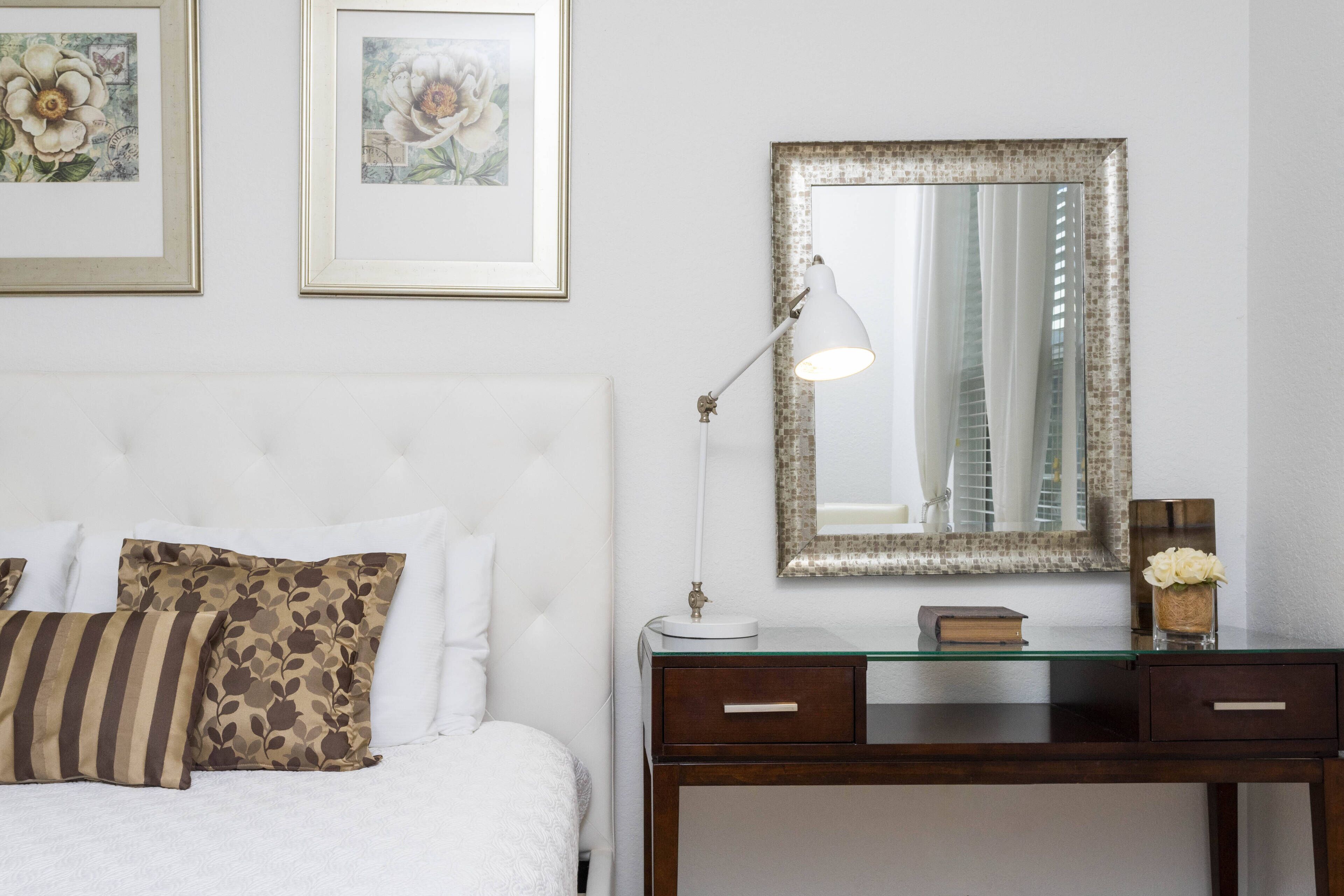 Un dormitorio ordenado con una cama, almohadas, una mesita de noche con una lámpara, un espejo y cuadros en la pared.