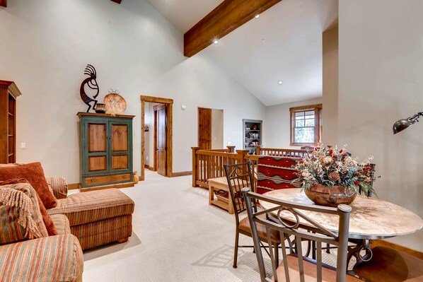 A spacious living area with a high ceiling, a dining table with chairs, a wooden cabinet, and a decorative wall piece.