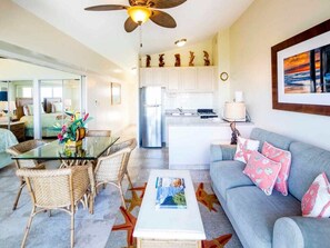 A modern living room with a glass dining table, wicker chairs, a grey sofa with patterned cushions, a coffee table, and a kitchen area with white cabinets and a stainless steel refrigerator. - Soak in Dazzling Pacific/Sunset Views, WiFi, Full Kitchen – Waikīkī Shore #PH09 (HONOLULU)