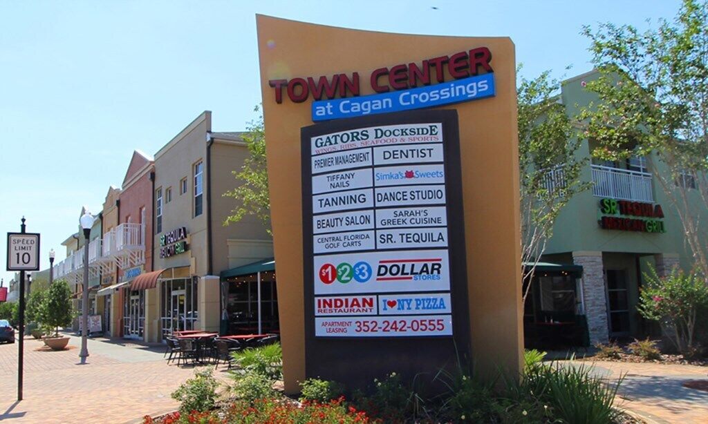Schild im Stadtzentrum mit einer Auflistung verschiedener Geschäfte, darunter Gators Dockside, ein Zahnarzt, Simkas Sweets, Solarium, Tanzstudio, Schönheitssalon, griechische Küche, Golfcarts Central Florida, SR. Tequila, Dollar Stores, ein indisches Restaurant und Wohnungsangebote.