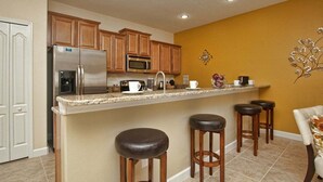 Cocina con armarios de madera, refrigerador de acero inoxidable, microondas, cafetera y una barra con taburete.