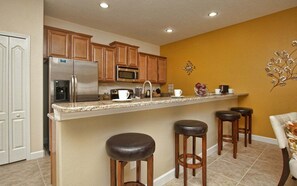 A kitchen with wooden cabinets, a stainless steel refrigerator, a microwave, a coffee maker, and a bar stool area. - Disney Pool Home | 2 King Bedrooms | Private Splash Pool | Paradise Palms Resort (Kissimmee)