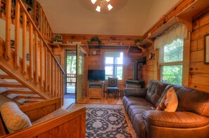 A cozy cabin interior with a wooden staircase, a leather sofa, a television, and a wood-burning stove. - 2BR cabin in Bryson City with hot tub, fireplace, WiFi, and mountain views. Free train and rafting (Bryson City)