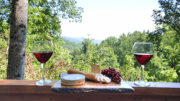Zwei Gläser Rotwein, ein Käselaib und Trauben auf einem Holztisch mit schöner Aussicht.