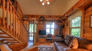 Ein gemütliches Blockhaus-Interieur mit einer Holztreppe, einem Ledersofa, einem Fernseher und einem Kaminofen.