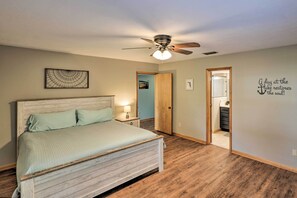 Un dormitorio con cama de madera, ventilador de techo, mesita de noche, puerta que da a otra habitación y una pared con una frase motivacional.
