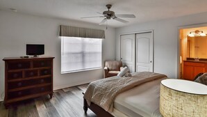 Ein Schlafzimmer mit einem Bett, einem Schminktisch, einem Fernseher, einem Deckenventilator, einem Fenster mit Jalousien und einer Tür.
