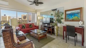 A spacious living room with a red sofa, a glass coffee table, wicker chairs, a TV, a desk, and a large window offering a city view. - Families Love the View and Fun Decor! Free WiFi, Full Kitchen–Waikīkī Shore #PH05 (HONOLULU)