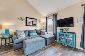 A living room with a grey sofa, a teal cabinet with books, a flat-screen TV, and a window with curtains.