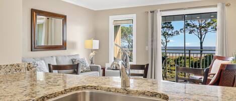 A modern kitchen with a granite countertop and stainless steel sink, a living area with a sofa and a lamp, and a balcony with a view of the ocean.