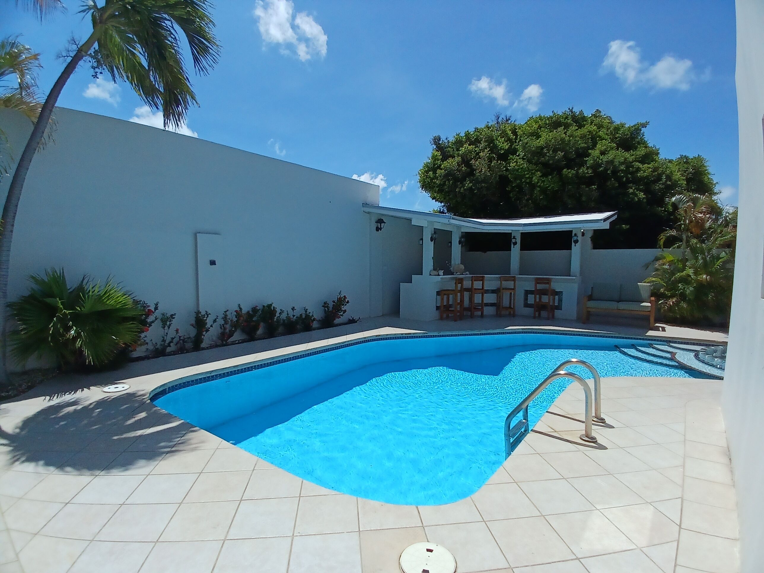 Una piscina con solárium de azulejos, una zona de estar cubierta con sillas y palmeras.