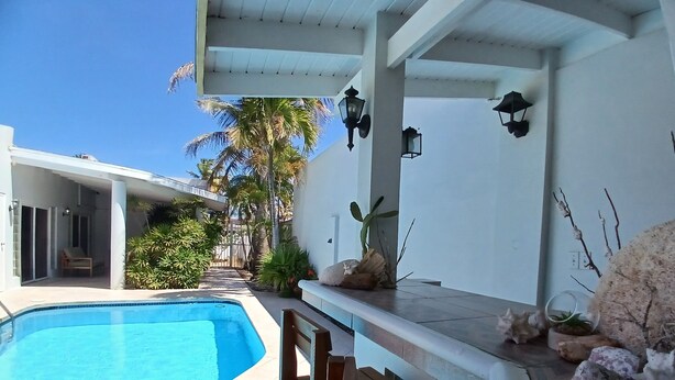 A pool area with a clear blue pool, a wooden bench, and a bar area with decorative stones. - Aruba private 3 bedroom holiday Villa, pool, walking distance to the beach (Noord)
