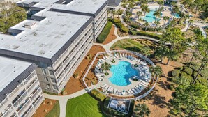 An aerial view of a hotel with multiple floors, a rooftop pool, and a landscaped pool area with lounge chairs and palm trees.