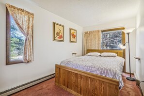 Une chambre à coucher avec un lit en bois, deux fenêtres avec des rideaux, une lampe sur pied et deux tableaux encadrés accrochés au mur.