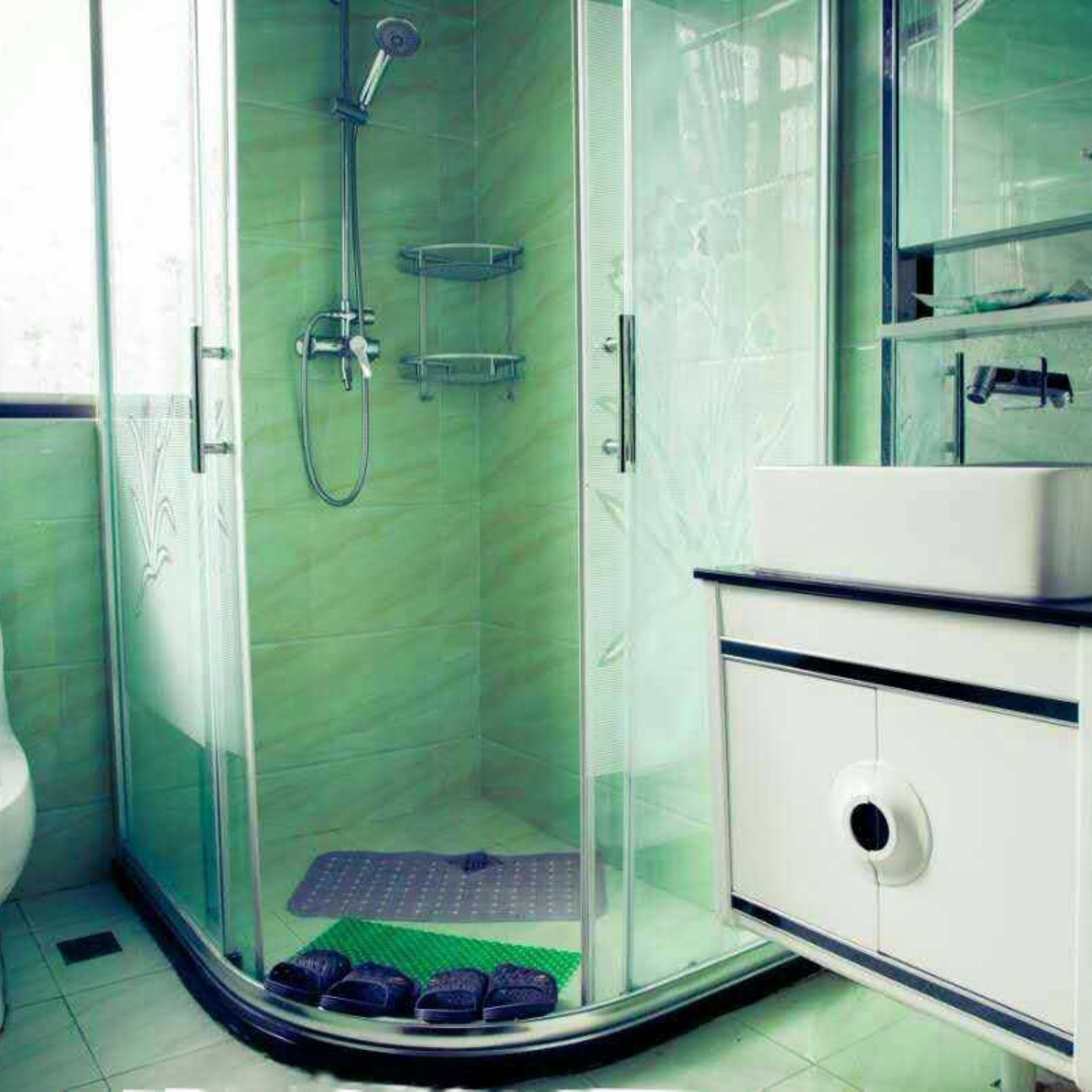 Un espace douche fermé par des parois de verre, doté d’un pommeau de douche à main, d’une étagère en verre et d’un lavabo blanc avec un miroir rond.