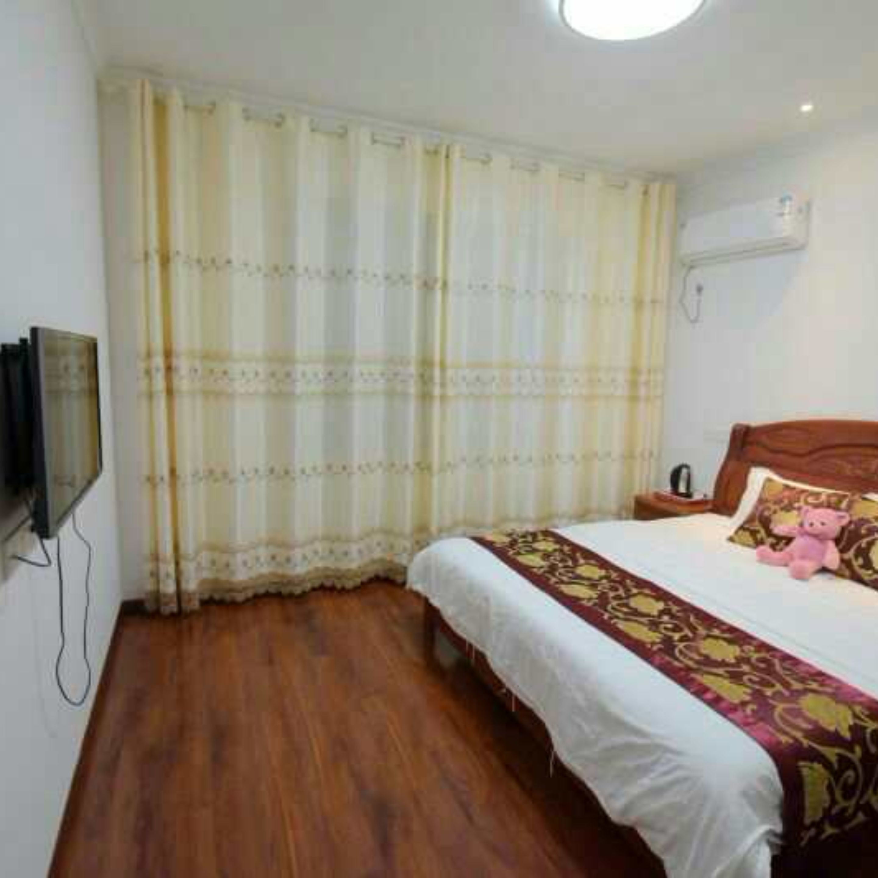 A bedroom with a wooden floor, a bed with a white and red bedspread, a pink teddy bear on the bed, and a television mounted on the wall.