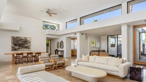 A spacious living area with a high ceiling, a dining table, comfortable seating, and large windows offering views of the outdoors.