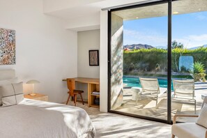 A modern bedroom with a large bed, a desk, and a view of an outdoor pool area. - Pet-Friendly Gated Home near Downtown (Palm Springs)