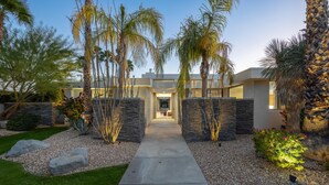 A modern house with a stone pathway leading to the entrance, surrounded by palm trees and a well-maintained garden.