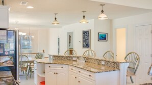 Una cocina con armarios blancos, encimeras de granito e iluminación colgante. Se observan sillas y una mesa de comedor.